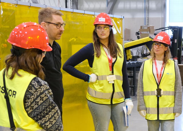 Three female visitors in red helmets listen while a male engineer from ESS is talking in a workshop
