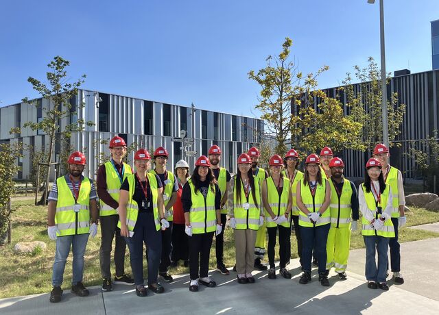 12 people stand in the sunshine wearing red helmets and yellow vests