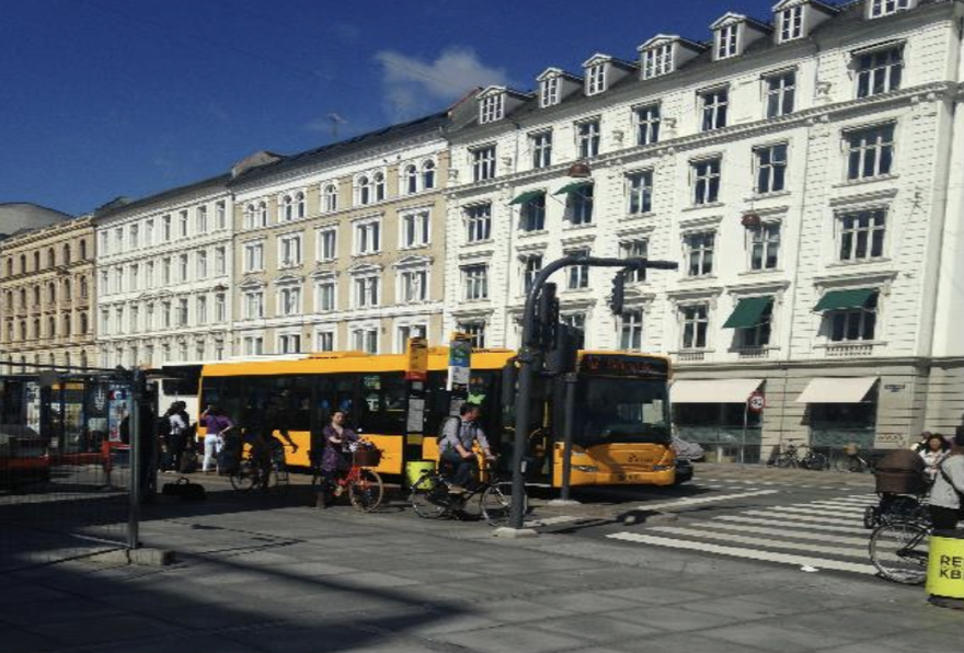 A yellow bus is parked near some traffic lights opposite a white building