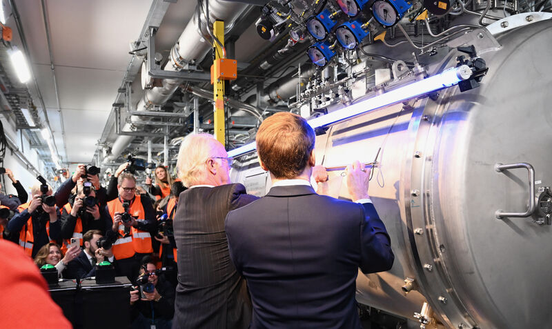 HRH The King of Sweden holds the left hand side of a metal plaque, while President Macron on his right holds the right hand side, and together they place it on the side of a shiny metallic cryomodule of the accelerator
