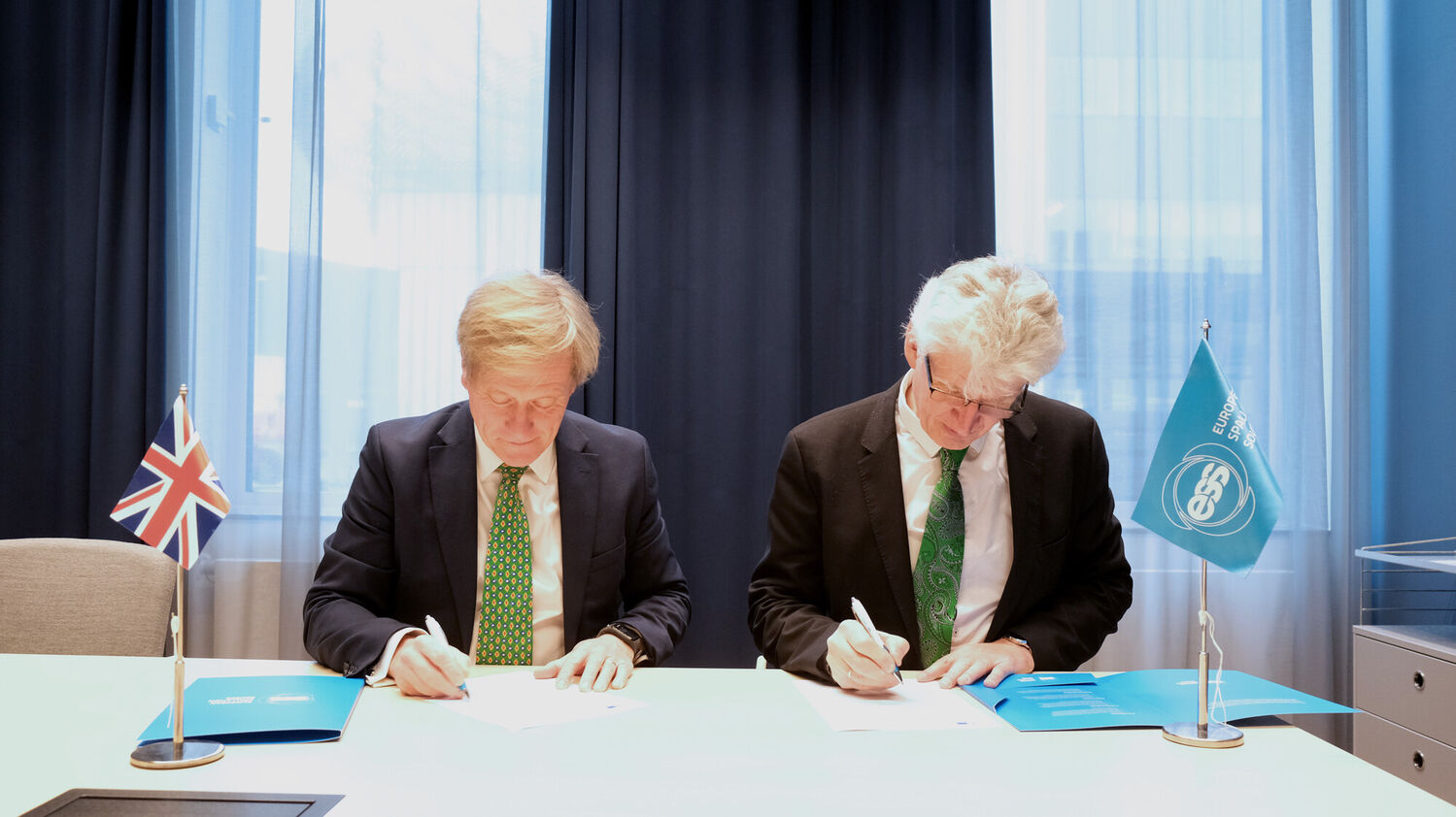 Two men in suits sit and sign documents with UK and ESS flags
